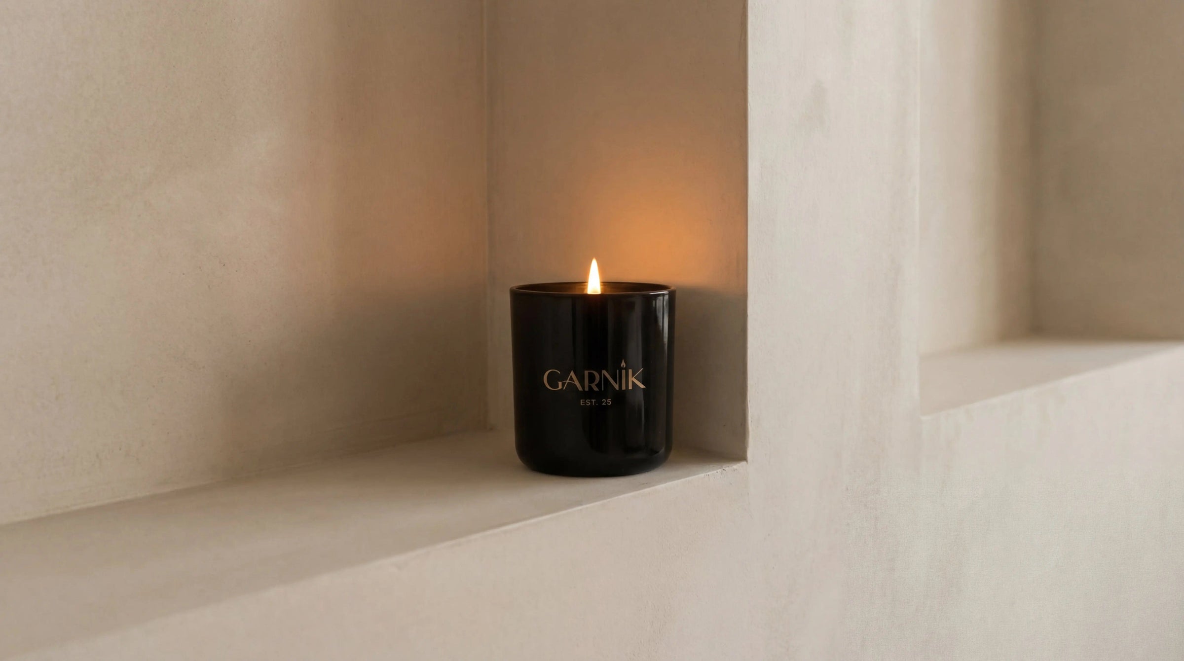 Garnik candle in a black jar placed on a white wall with a soft glow.